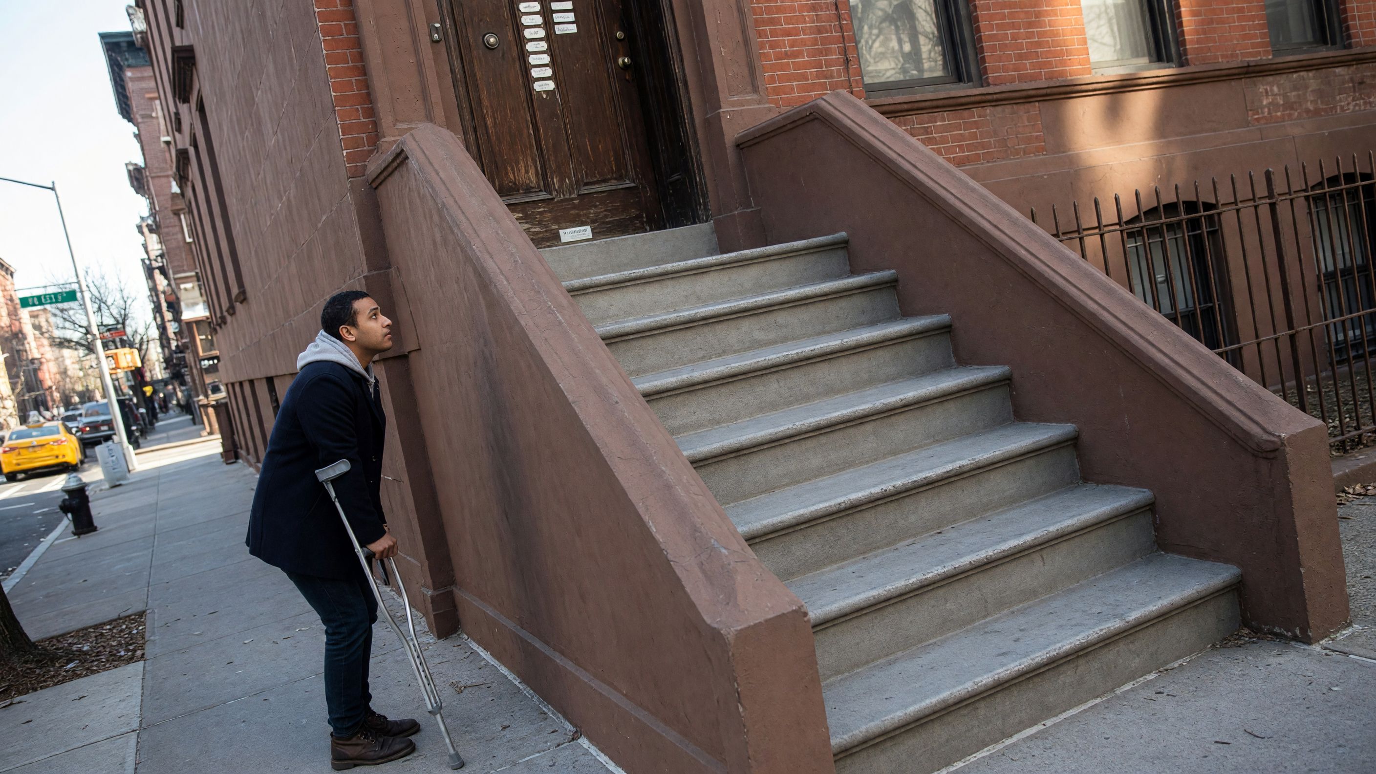 A man with a crutch looks up a stoop of stairs to a closed entrance. There is no ramp.